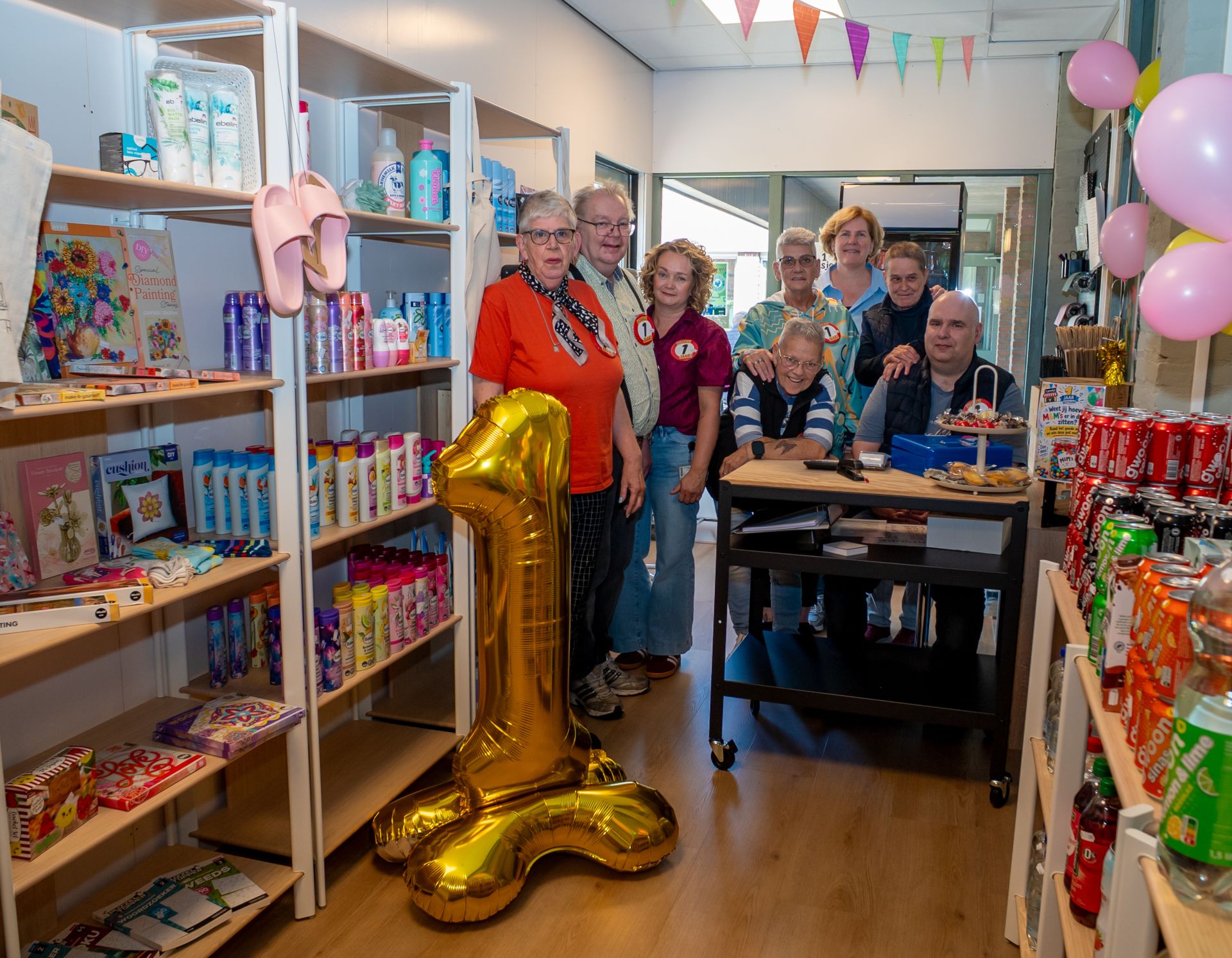 Een groep mensen viert de eerste verjaardag van een winkel met decoraties en feestelijke ballonnen.