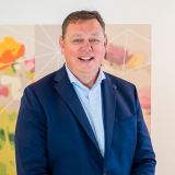 Een man in een blauw pak staat voor een kleurrijk bloemenkunstwerk.