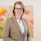 Een glimlachende vrouw in een beige jasje staat voor een kleurrijk bloemenschilderij.
