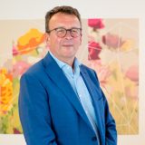 Een man in een blauw pak staat voor een kleurrijke bloemenafbeelding.